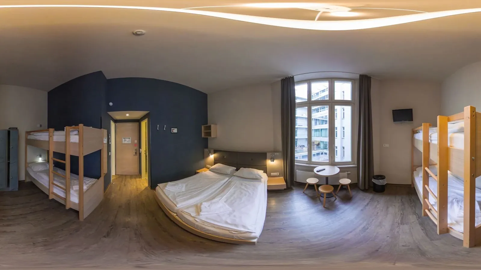 Rundumansicht eines Hotelzimmers mit Doppelbett, zwei Etagenbetten, kleinem runden Tisch mit Hockern und großem Sprossenfenster