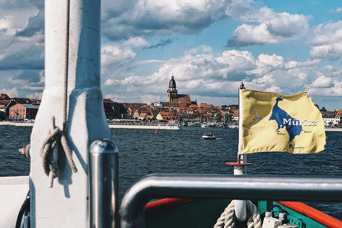 Blick vom Ausflugsschiff über die Müritz auf Waren (Müritz) mit der Marienkirche, an der Reling weht eine gelbe Müritz-Flagge