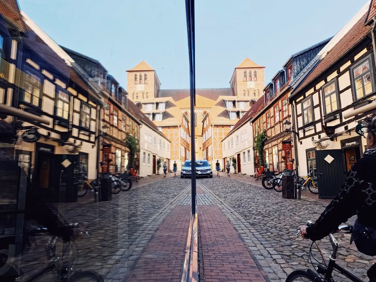 Altstadtgasse mit Fachwerkhäusern und zwei Backsteintürmen in Waren (Müritz), symmetrisch in einer Schaufensterscheibe gespiegelt, rechts ein Radfahrer